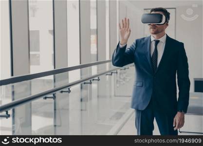So real! Young businessman with VR glasses on head standing in office, excited office worker dressed in formal clothes managing business project through virtual reality, touching object with hand. Excited office worker dressed in formal clothes managing business project through virtual reality