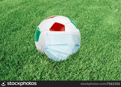 Soccer ball in italian colors with mask on green field. Virus threatened championship concept. Virus in footbal