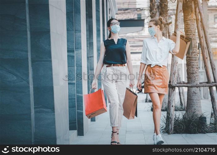 social distancing new normal concept.portrait of two happy girls walking together wearing medical face masks, walking outdoors in the evening near mall after covid epidemic , holding shopping bags.