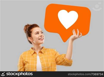 social media marketing, internet and people concept - smiling red haired teenage girl in checkered shirt holding heart icon over grey background. red haired teenage girl holding speech bubble