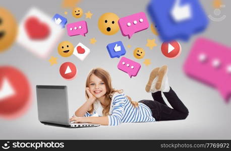 social media, technology and blogging concept - smiling little student girl with laptop computer lying on floor with internet icons over grey background. smiling girl with laptop and internet icons