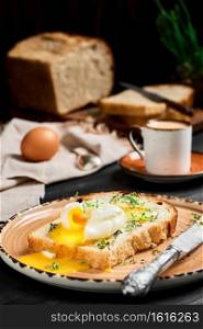 Soft-boiled  poached  egg on slice of bread covered with butter cream and herbs, on clay plate on black wooden table. Espresso coffee and loaf of sliced bread on blurred background. Breakfast idea
