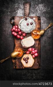 Soft cheese selection with grape and Honey mustard sauce on rustic cutting board on dark background, top view