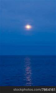 Soft moon in clouds and haze reflected in water on blue evening sky.