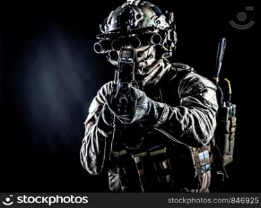 Soldier of special forces in camo combat uniform, load carrier, helmet, equipped night sight goggles, tactical radio headset, aiming assault rifle with collimator sight in camera, low key studio shoot. Army soldier aiming rifle in camera in darkness