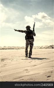 Soldier of special forces, infantry rifleman in military ammunition walking in desert and pointing on horizon. Military reconnaissance team leader managing dead ground observation with combat patrol. Army commando soldier observing sandy desert area