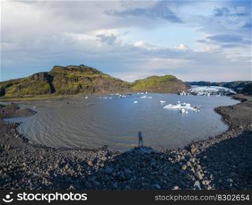 Solheimajokull glacier, Iceland. Its tongue slides from the volcano Katla. Glacial lake lagoon with ice blocks and surrounding mountains. Hill top and unrecognizable photographer shadow in water.
