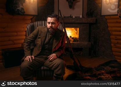 Solid male hunter in traditional hunting clothes sitting in a chair against burning fireplace. Retro style, vintage rifle. Stuffed wild animals, bear skin on background