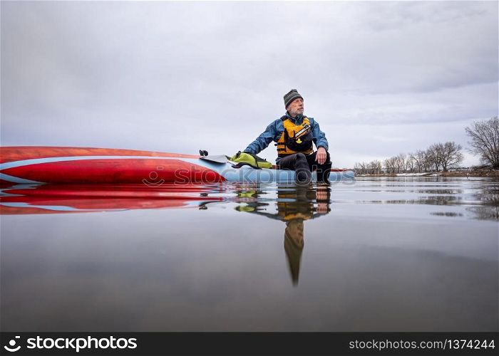 solo lake paddling as social distancing recreation during coronavirus pandemic, a senior male paddler has a moment of reflection when sitting on his paddleboard in the middle of calm lake