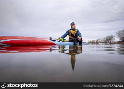 solo lake paddling as social distancing recreation during coronavirus pandemic, a senior male paddler has a moment of reflection when sitting on his paddleboard in the middle of calm lake