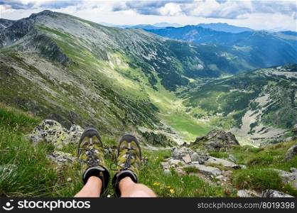 Somebody is sitting at the edge of cliff in mountains, showing his feet wearing hiking shoes. First person shoot. Retezat area, Carpathians, Romania
