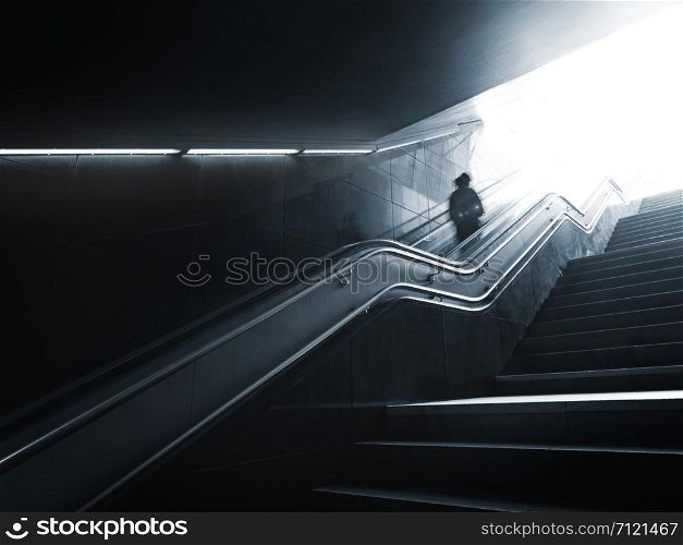 Someone on escalator is moving up to bright daylight from darkness and electric light of underground, motion blur and flares, monochrome image