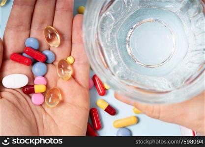 Someones hand holding colorful pills with glass of clear water over blue background. Medical pharmacy concept.. Pile of pills