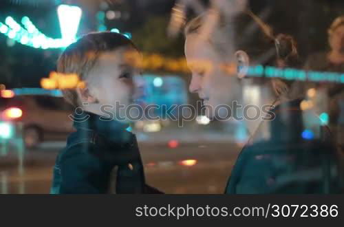 Son and mother in the busy street at night. Excited child talking to the mom, she embracing and kissing him. View through the glass