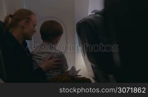 Son and mother traveling by air. Child sitting on moms lap and opening illuminator blind, they looking out it together