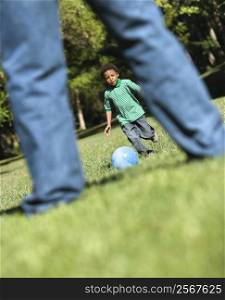 Son running and kicking ball towards father in park.