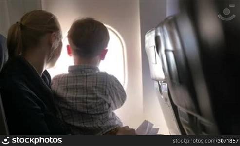 Son sitting on mothers lap in plane and they enjoying view from illuminator, mom kissing the boy and telling something to him