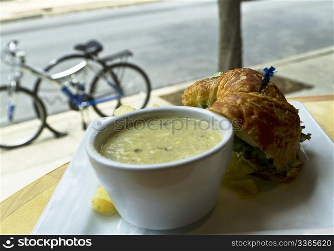 Soup and sandwich stop with bicycles parked outside and visible through restaurant window; cup of vegetable chowder with croissant bread, vegetable sandwich, edge of potato chip showing.