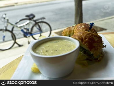 Soup and sandwich stop with bicycles parked outside and visible through restaurant window; cup of vegetable chowder with croissant bread, vegetable sandwich, edge of potato chip showing.