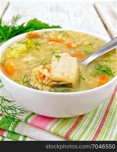 Soup fish kulesh with millet, potatoes and carrots and spoon in a white bowl on a napkin, parsley, dill on a wooden board background