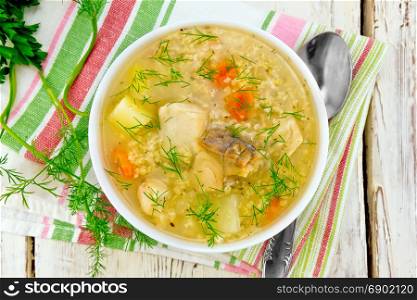 Soup fish kulesh with millet, potatoes and carrots in a white bowl on a napkin, parsley, dill on a wooden board background on top