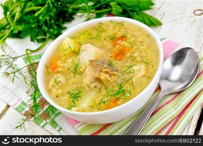Soup fish kulesh with millet, potatoes and carrots in a white bowl on a napkin, parsley, dill on a wooden board background
