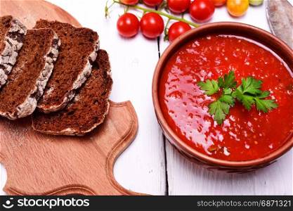 Soup of fresh red tomato gazpacho in a ceramic brown plate on a white table, next to sliced pieces of rye bread on a kitchen board, top view