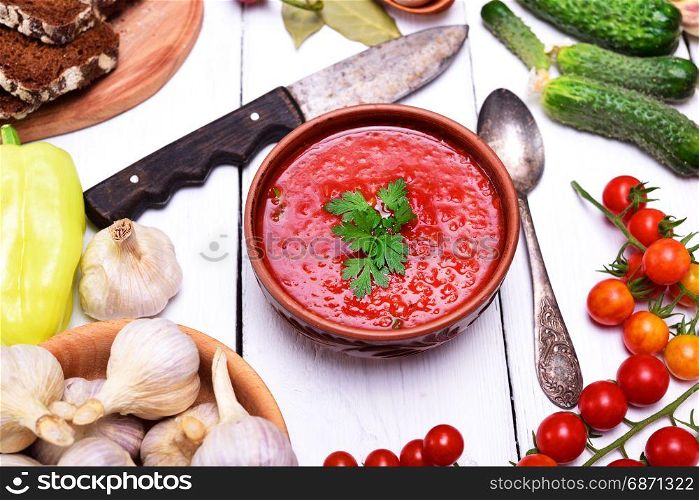 Soup of fresh vegetables gazpacho in a ceramic brown plate on a white table, around the ingredients for cooking on a prescription