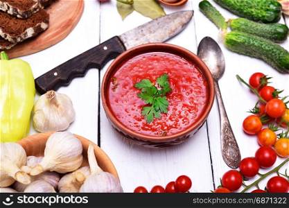 Soup of fresh vegetables gazpacho in a ceramic brown plate on a white table, around the ingredients for cooking on a prescription