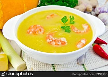 Soup-puree pumpkin with shrimp and parsley in a white bowl on a kitchen towel with a spoon, squash, parsley, ginger, hot red pepper pods, leek on a wooden boards background