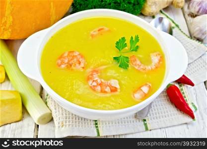Soup-puree pumpkin with shrimp and parsley in a white bowl with a spoon on the towel, pumpkin, parsley, ginger, hot red pepper pods, leek on a wooden boards background