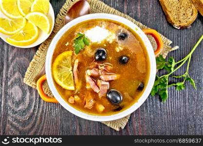 Soup saltwort with lemon, meat, pickles, tomato sauce olives in a bowl on a sacking, bread on a background of a dark wooden board from above