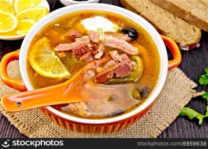 Soup saltwort with lemon, meat, pickles, tomato sauce olives, spoon in a bowl on a napkin of burlap on the background of a wooden board. Solyanka with lemon and black olives in bowl on board