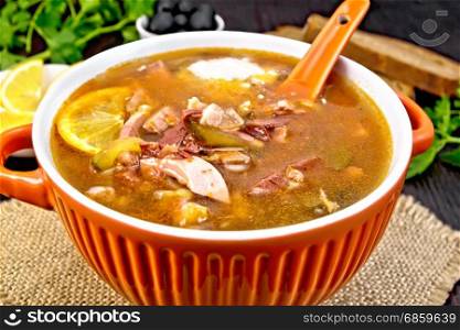 Soup saltwort with lemon, meat, pickles, tomato sauce olives, spoon in a bowl on a sacking on a background of a dark wooden board