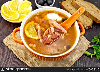 Soup saltwort with lemon, meat, pickles, tomato sauce olives, spoon in a bowl on a napkin of burlap on the background of a dark wooden board. Solyanka with spoon in bowl on dark board