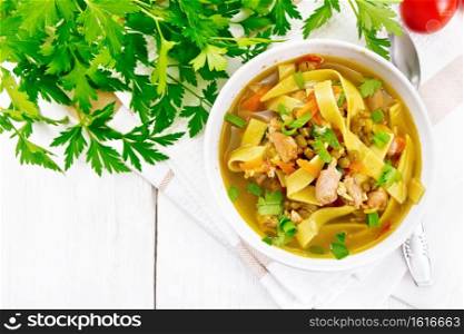 Soup with meat, tomatoes, vegetables, mung bean lentils and noodles in a bowl on napkin, parsley and a spoon on the background of light wooden board from above