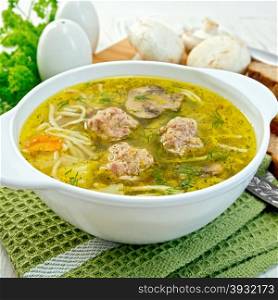 Soup with meatballs, noodles and mushrooms in a white bowl on a napkin, parsley on the background light wooden boards