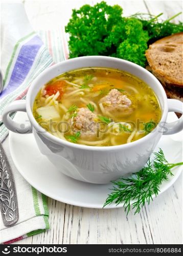 Soup with meatballs, noodles and vegetables in a white bowl and saucer, dill, bread, napkin and spoon on a wooden boards background