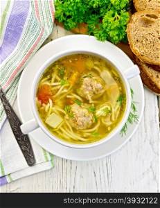 Soup with meatballs, noodles and vegetables in a white bowl, bread, napkin and spoon on the background light wooden boards on top