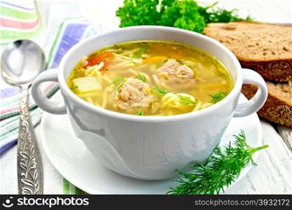 Soup with meatballs, noodles and vegetables in a white bowl, bread, napkin and spoon on the background light wooden boards
