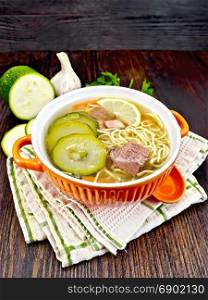 Soup with zucchini, beef, ham, lemon and noodles in a bowl, parsley and dill on a napkin on a wooden board background