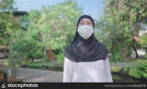 South east Asian Muslim female wear protective face mask. Hijab woman answering phone during a walk inside public park, phone call, Coronavirus COVID-19 pandemic lifestyle in Islamic country culture