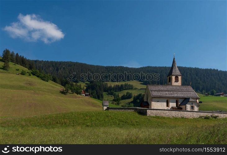 South Tyrolean mountain church under a blue sky with a single white cloud, mountain landscape with meadows and pines