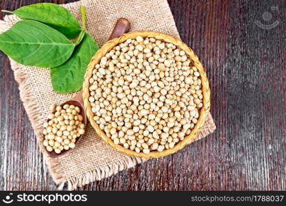 Soy beans in wicker bowl and spoon, green leaf on burlap on wooden board background from above