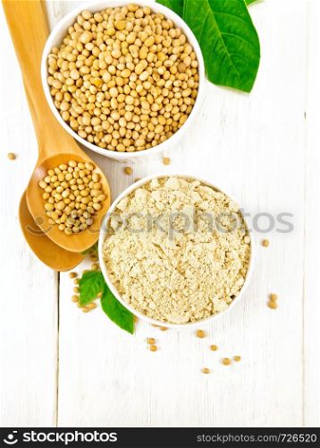 Soy flour and soybeans in two bowls, spoons and green leaves against a white wooden board from above