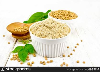 Soy flour and soybeans in two bowls, spoons and green soybean leaves against a white wooden board