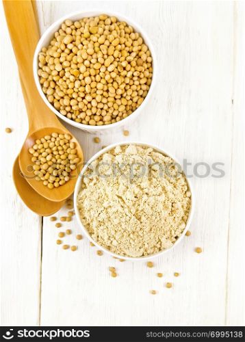 Soy flour and soybeans in two white bowls, spoons on a wooden board background from above