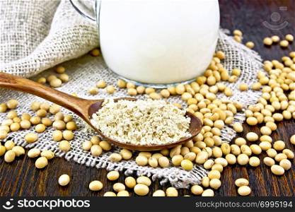 Soy flour in a spoon and milk in a glass jug, soybeans on napkin of burlap on wooden board background