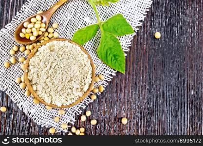 Soy flour in the bowl, soybeans in a spoon and on a napkin of burlap sacking, green leaf on the background of a wooden board on top