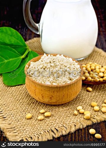 Soy flour in the bowl, soybeans in a spoon and on a napkin of burlap, milk in a jug, green soya leaf against a dark wooden board